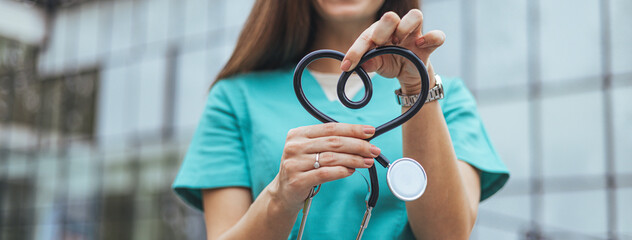A caucasian female nurse in teal scrubs holds a stethoscope, the tubing shaped like a heart,...