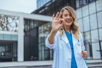 A joyful female healthcare worker extends a welcoming hand, dressed professionally in a lab coat, set against the modern facade of a healthcare facility.