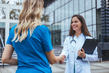 Two Caucasian female healthcare workers collaborate outside a medical facility; one in a lab coat...