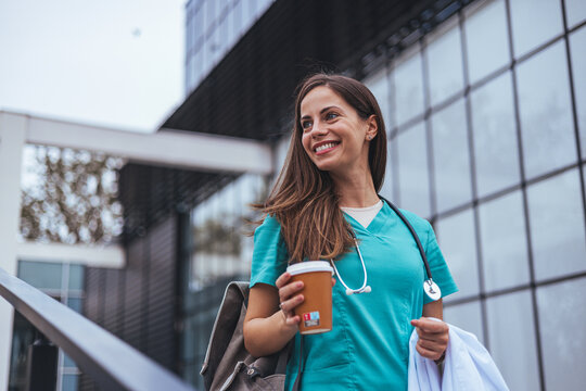 A cheerful Caucasian female nurse, clad in scrubs with a stethoscope, relishes a break while holding a coffee cup, standing before a medical facility.