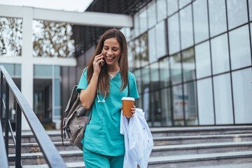 A cheerful Caucasian woman in scrubs engages in a phone conversation while holding a coffee cup,...