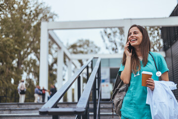 A Caucasian woman in a teal scrub holds a coffee cup and chats on her mobile phone, radiating joy...