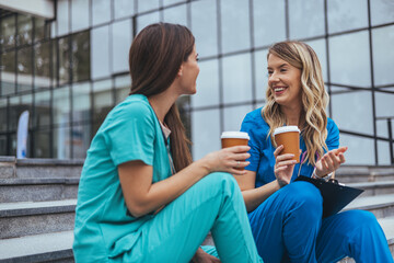 Two smiling Caucasian female nurses in scrubs relax with coffee on steps outside a medical...