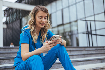 A cheerful female healthcare worker in blue scrubs sits on steps outside a building, browsing her smartphone during a break.