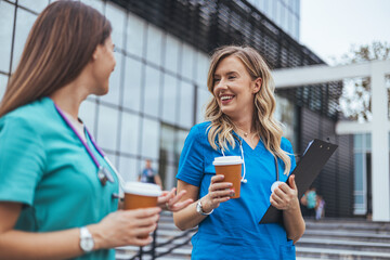 Two smiling Caucasian female nurses in scrubs relax outside a medical facility, holding coffee cups...