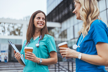 Two smiling female nurses in blue scrubs relax with coffee cups during a break outside a medical...