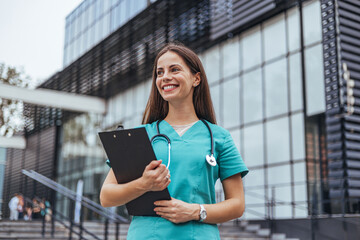 A joyful Caucasian female nurse stands confidently outside a hospital, holding a clipboard while...