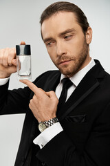 A dapper bearded businessman in a suit confidently holding a bottle of perfume against a grey studio backdrop.