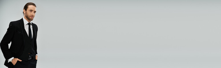 Handsome, bearded businessman in suit and tie stands confidently with hands in pockets on a grey studio background.