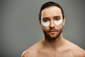 A handsome, shirtless man with a beard confidently wears eye patches in a striking pose on a grey...