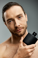 A handsome shirtless man with a beard holds perfume bottle against a grey background in a studio setting.