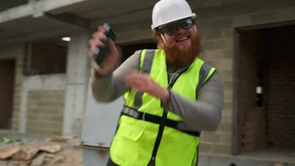 Bearded male builder dancing jubilantly waving his arms in sunglasses and helmet walking through the construction site different successfully completed project and the end of the working day.