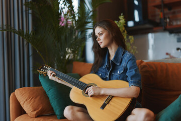 Woman playing acoustic guitar on couch by window in natural light ambiance with gentle artistic...