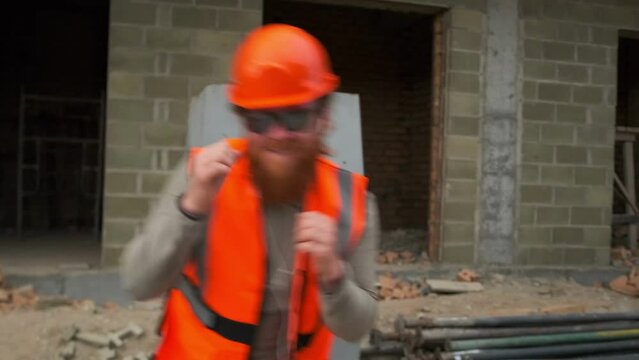 Young funny male worker in orange protective suit makes boxing dance moves and dancing at construction site. Successful deal, salary increase, successful project.