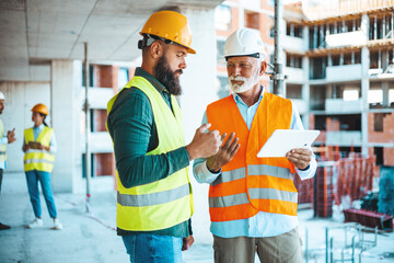 A construction worker and a site manager in safety gear engage in a discussion over a digital tablet, with team members working in the background at the bustling site.