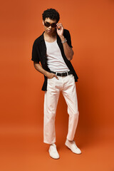 Handsome man in white pants and black shirt poses on vibrant orange backdrop.