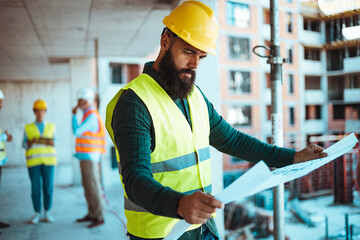 A bearded man in a safety vest and hard hat reviews construction plans at a busy site, with diverse...