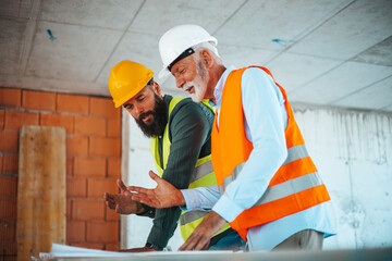 Two male construction site managers, with diverse backgrounds, are analyzing blueprints at a building project.