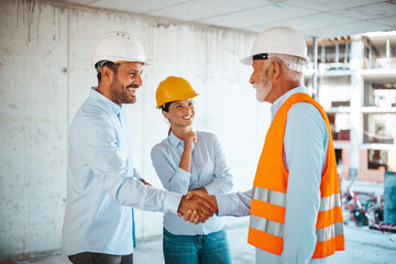 Two male construction site managers in hard hats and safety vests shake hands while a female...