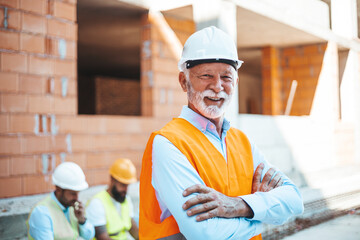 A senior male construction site manager, dressed in a white helmet and high-visibility vest, smiles...