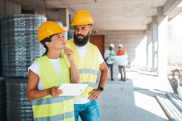 A male and a female worker in safety gear discuss project details, with clipboard in hand, at a...