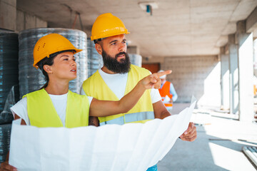 A male and a female construction worker in safety gear examine project plans together at a building...