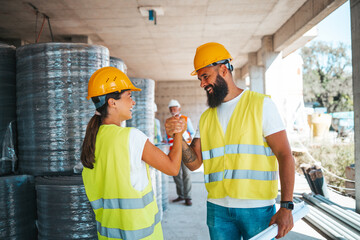 A smiling male and female construction worker shake hands on a busy construction site, attired in...