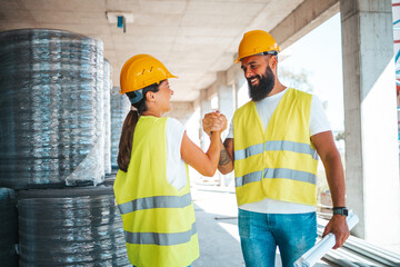 A male and female worker in safety vests and helmets engage in a firm handshake, exemplifying teamwork on a construction site.