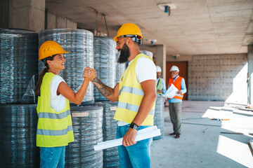 A male and female in safety gear engage in a firm handshake at a bustling construction site, with...