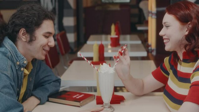 Side shot of happy Caucasian couple sipping milkshake from same glass using straws and holding hands, spending time together in cozy retro American diner