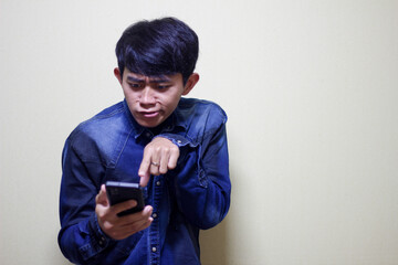 A young man with a furrowed brow and a puzzled expression is deeply engrossed in his smartphone,...