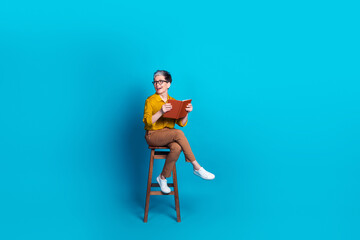 Full length photo of adorable good mood lady dressed shirt eyewear sitting stool read novel empty space isolated blue color background