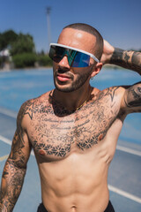 Chico joven musculoso y tatuado con gafas deportivas posando en pista de atletismo en pleno dia