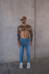 Chico joven musculoso y tatuado posando con vaqueros frente a pared
