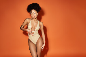 African American woman in trendy swimsuit poses against vivid orange backdrop.