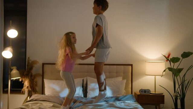 Joyful family hyperactive Caucasian children little boy girl bouncing holding hands play together jump bed son daughter bad behavior sleeping night kids sleepover inside bedroom illuminated apartment