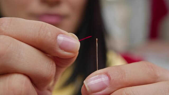 Seamstress carefully threads a thin red thread through the eye of the needle. After several attempts, she manages to thread it successfully. Close up. Slow motion.