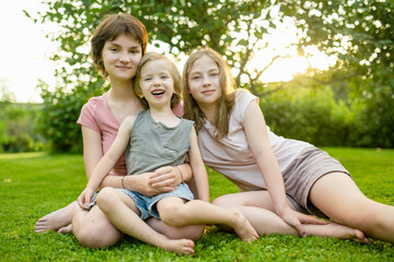 Fototapeta premium Cute big sisters cuddling with their toddler brother. Adorable teenage girls hugging their small sibling. Children exploring nature.