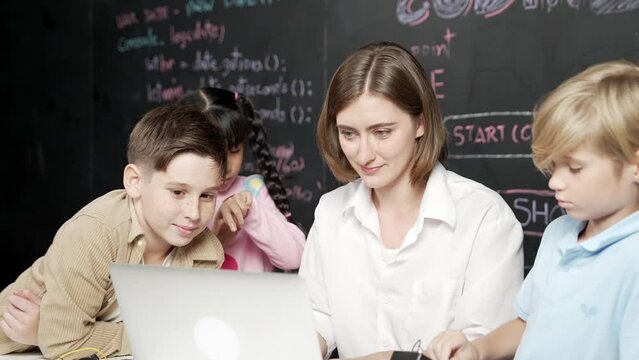 Caucasian teacher using laptop and explain about engineering system while diverse student holding electronic equipment in front of blackboard with prompt or code written in STEM class. Erudition.