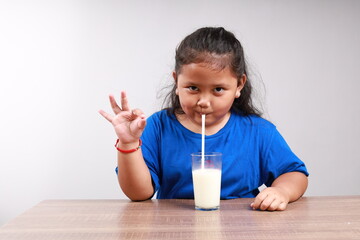 Enjoyed Adorable asian little girl drinking a glass of milk using straw while making okay hand...