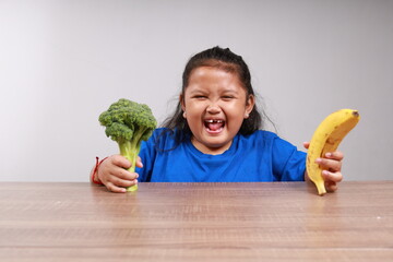 Adorable asian little girl feels exciting while holding broccoli and a banana. Healthy food concept
