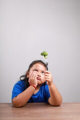 Unhappy asian little girl refusing to eat vegetables at table in room