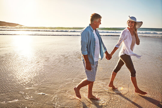 Happy couple, walk and holding hands on beach for getaway on island or coast for vacation, travel and holiday. Woman, man and relationship for love or marriage anniversary by ocean at sunrise in Bali
