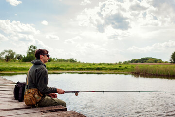 Young man fishing wearing military uniform. Guy sitting on bridge across river holding rod....