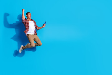 Fototapeta premium Full length photo of ecstatic guy wear stylish t-shirt hold smartphone jump win lottery look empty space isolated on blue color background