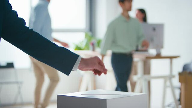 Close up of hand putting slip of paper with UK Union Jack flag on it into ballot box at polling station for United Kingdom election with voters - shot in slow motion