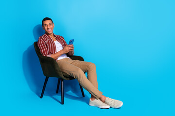 Full length photo of cheerful guy wear striped stylish t-shirt sit on armchair holding smartphone isolated on blue color background