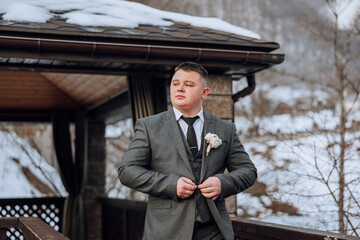 A man in a suit and tie is standing on a bridge, adjusting his tie. The scene is set in a snowy environment, with a building in the background. The man is getting ready for a formal event