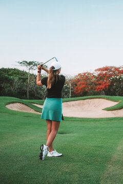 View from the back of a young woman hitting the ball with a golf club and looking into the distance to see if she hit the hole. Professional golf at a country club. Summer luxury sport.