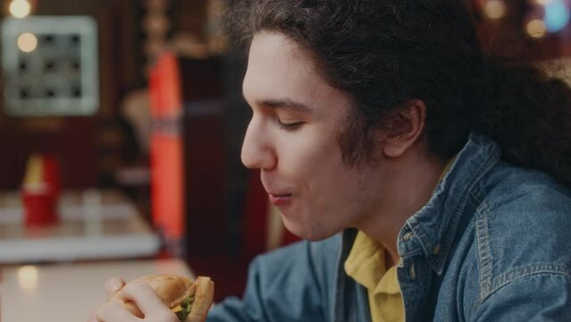 Side closeup of happy Caucasian guy in denim jacket and with long hair devouring burger in retro style diner and enjoying life